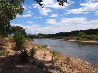 [White clouds scattered across a pale blue sky top the scene of the shallow Colorado River flowing into the foreground. Both banks of the river although this photo was taken from the left bank.]